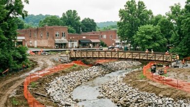 A newly excavated creek channel with rock banks and a pedestrian bridge under construction beside a small downtown
