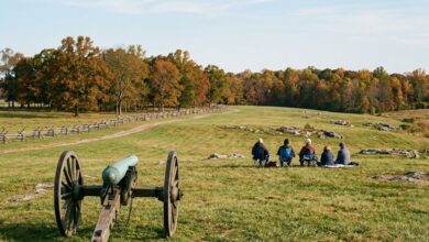 A bronze field cannon on a wooden carriage sits on a battlefield park lawn as visitors watch from folding chairs, with split-rail fencing and autumn hardwoods behind (illustrative)